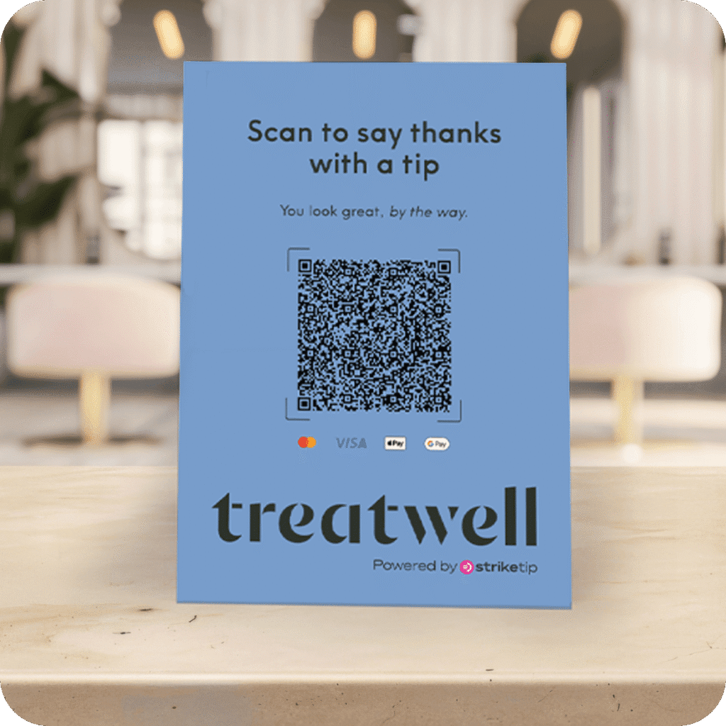 A blue tabletop display card stands on a wooden counter inside a blurred salon setting. The card reads "Scan to say thanks with a tip" and "You look great, by the way." above a large QR code. Below the QR code are the logos for Mastercard, Visa, Apple Pay, and Google Pay, followed by a large "Treatwell" logo and the text "Powered by striketip".